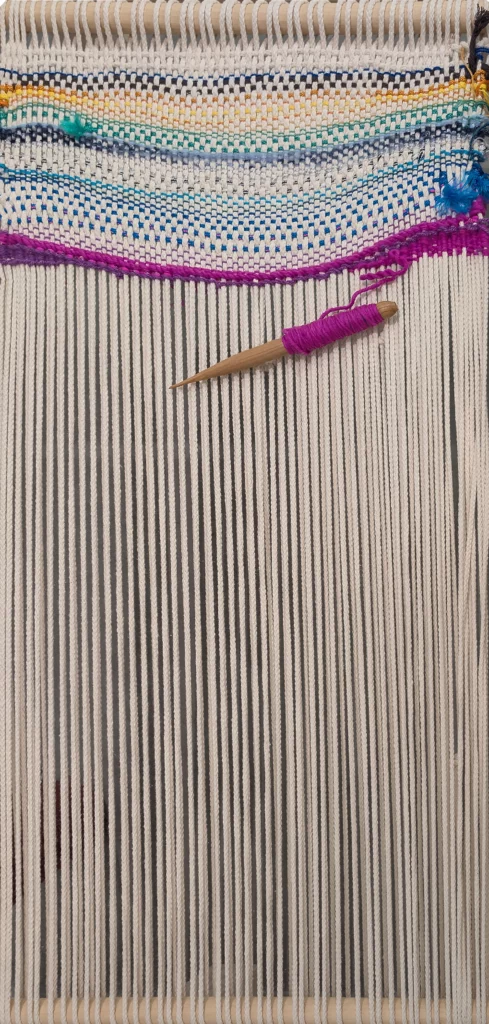 Detail of the loom used to create Otobong Nkanga’s Loom 05, with white threads interwoven with blue, green and purple lines, and a wooden shuttle wrapped in purple yarn.