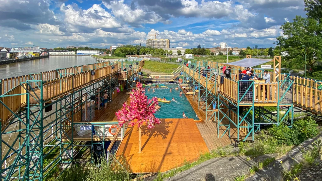 Piscine extérieure temporaire entourée de structures en bois et en métal, installée au bord du canal à Bruxelles dans le cadre de Right to the City, Right to the Future.