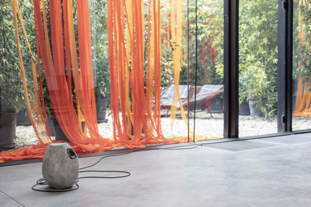 Vue de l’installation Blindsight de Manon de Boer, avec un haut-parleur intégré dans une forme ovoïde posée au sol devant de longues bandes de tissu orange suspendues.